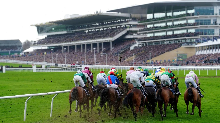Racehorses jumping a fence in front of the grandstand at a UK racecourse